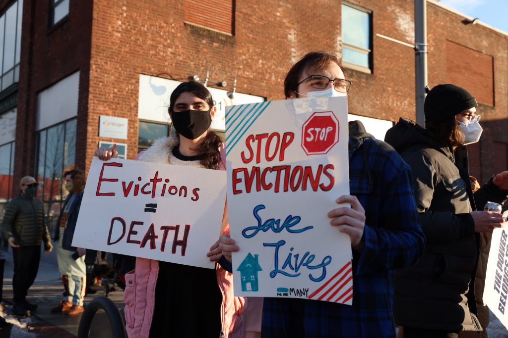 Two people holding signs. One reads `Evictions = Death` the other says `Stop Evictions Save Lives