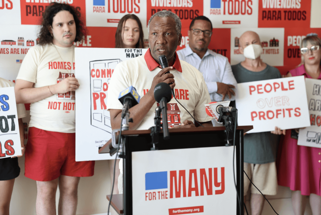 Councilman Anthony Grice speaks in to a microphone at a press conference. Behind him, people stand holding signs calling for short-term rental regulation.