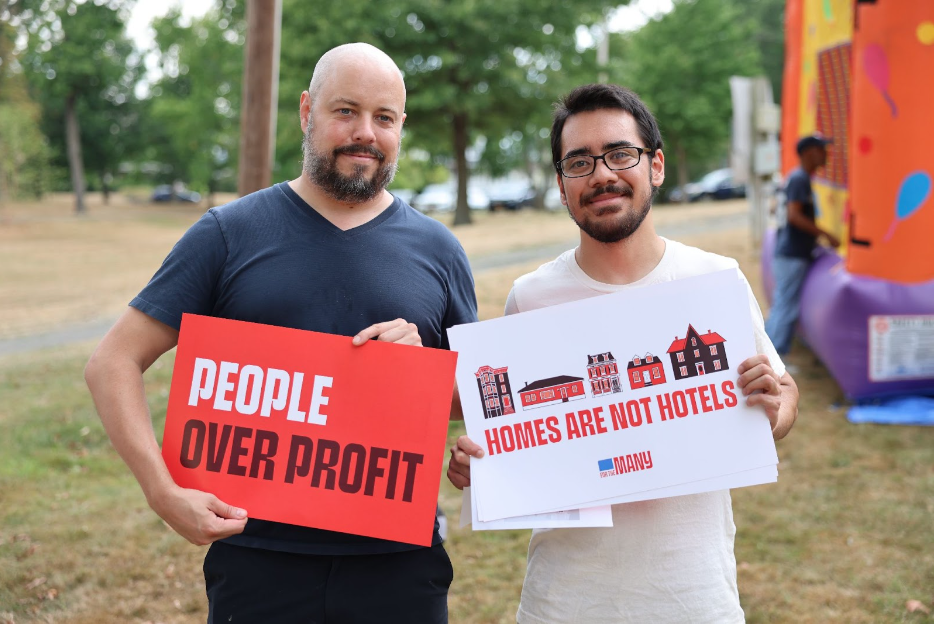Two men hold signs that say "People Over Profit" and "Homes Are Not Hotels" 